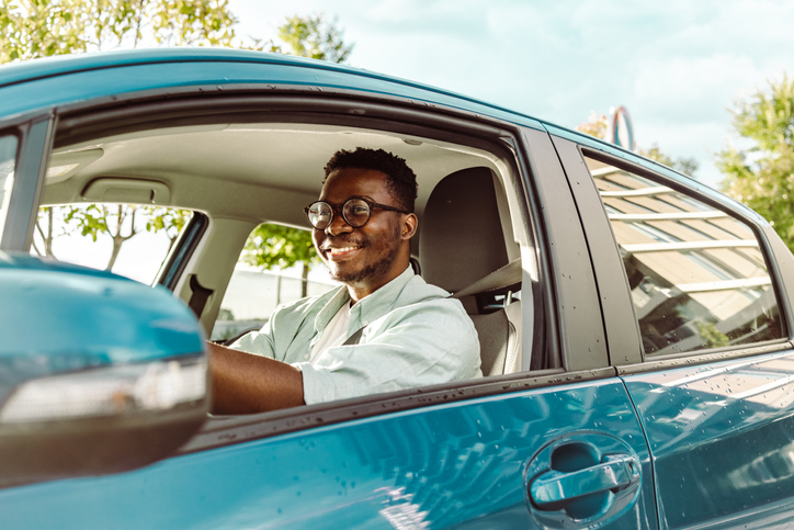 A male with dark skin and hair, wearing a light-colored shirt smiles as he sits in the driver's seat of his new, blue vehicle.
