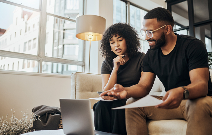 A man and woman sit on a sofa as they lean over an open computer and review financial documents.