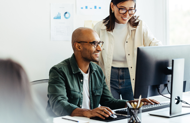 A male and female business professional smile as they look at the screen of a computer.