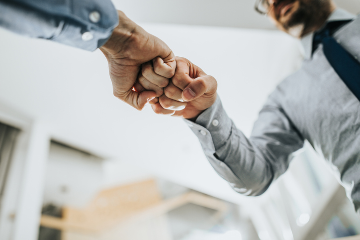 Two people in business casual attire fist-bump in an office.