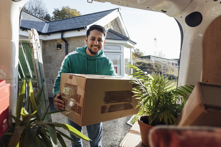 A smiling mid-adult man wearing casual clothing unloading boxes and furniture from the rear of a moving van, ready to move in to his new bungalow.