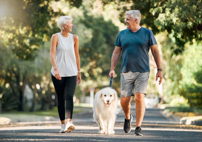Retirement age couple smiling while walking on a path with a dog.