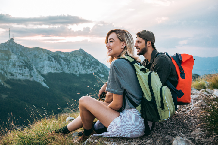 A young male and female sit at the top of a mountain hike as they enjoy the view.