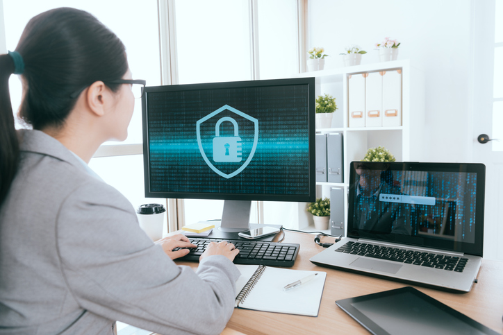 Woman sitting in front of a laptop and desktop screen with a security lock on it.