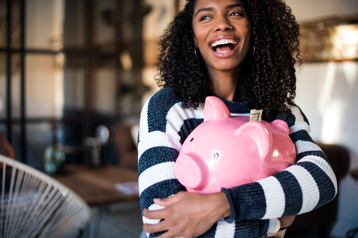 Young black woman smiling while holding a pink piggy bank with money sticking out of the top.
