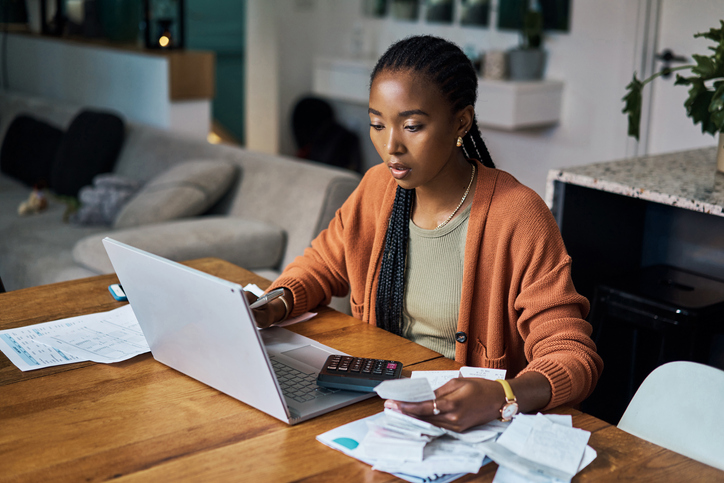 Young black woman sitting at a table in front of a laptop while going through bills.