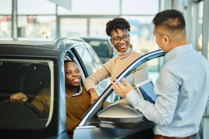 Happy black couple testing a car while talking to a car salesperson.