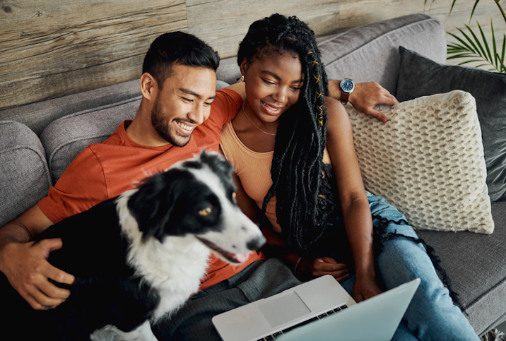Young couple sitting in front of a laptop smiling with their dog.