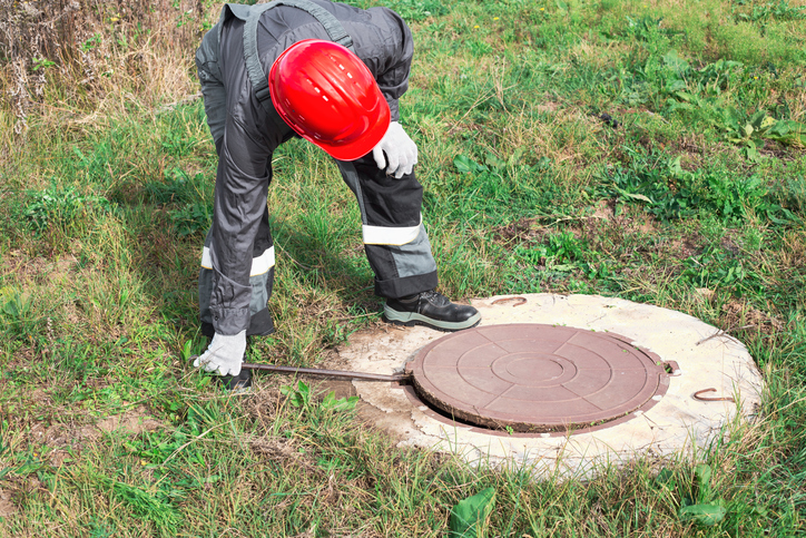 A worker in overalls and a helmet opens a manhole with a crowbar.