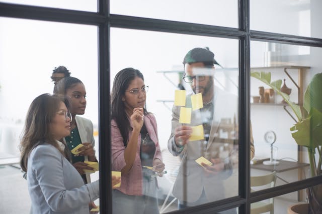 Four people in business casual attire looking at a board with sticky notes on it.