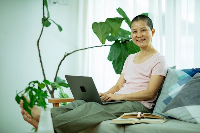 Middle-aged Asian woman sitting on a couch with a laptop while smiling.