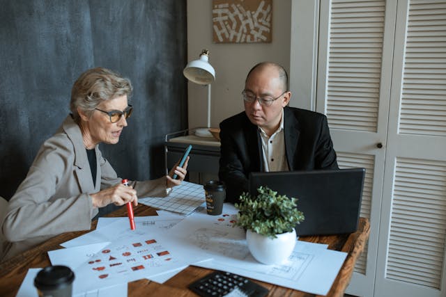 Older woman sitting at a desk and talking to a man while looking over papers.