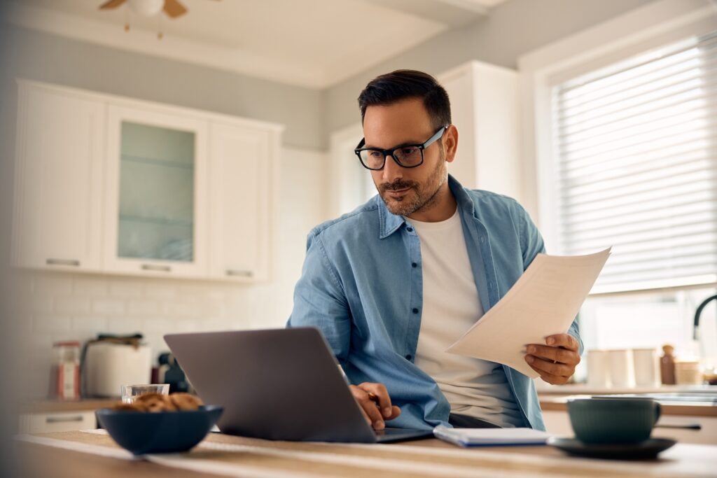 Man sitting in the kitchen and going over finances on a laptop while holding papers.