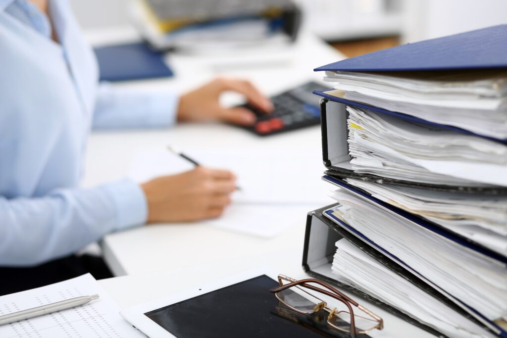 Binders with papers in the foreground with a businesswoman in the background working.