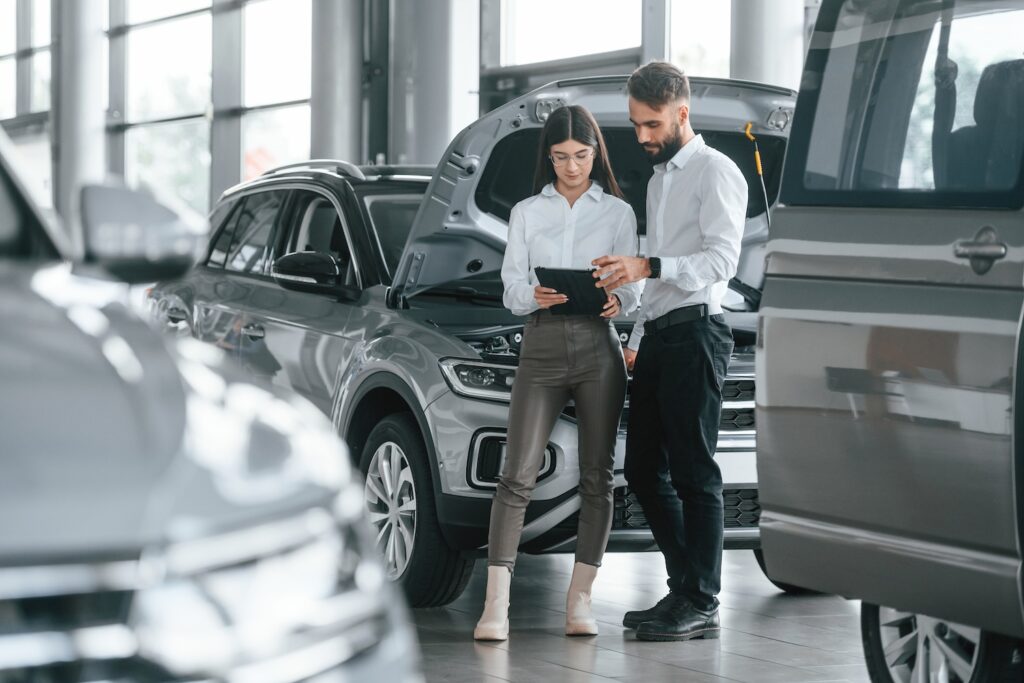 Two people standing together at a car dealership looking at a tablet.
