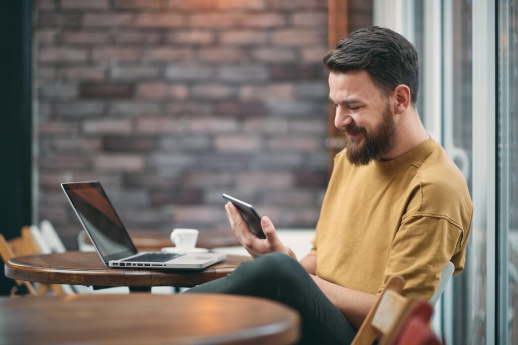 Man sitting at a table looking at his phone while also on his computer.