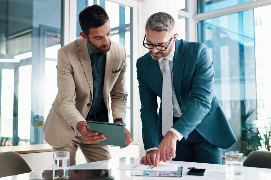 Two men discussing numbers while looking at a tablet and print out.