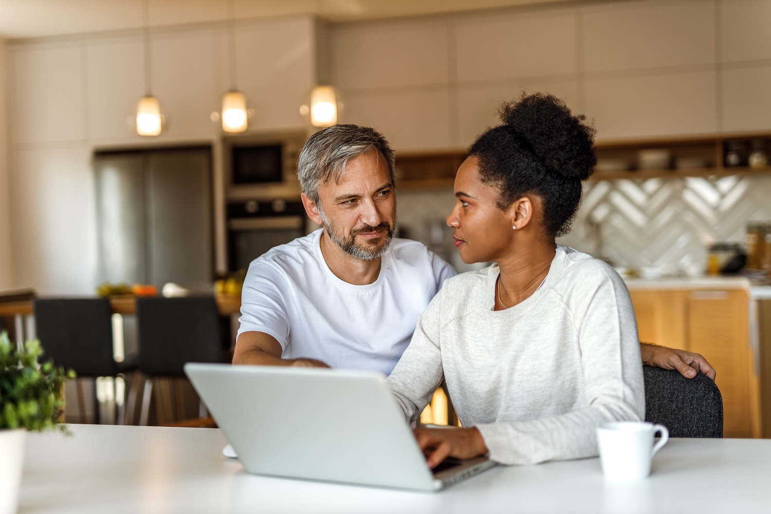 A man with graying hair and a woman sitting together at a kitchen table looking at a laptop.