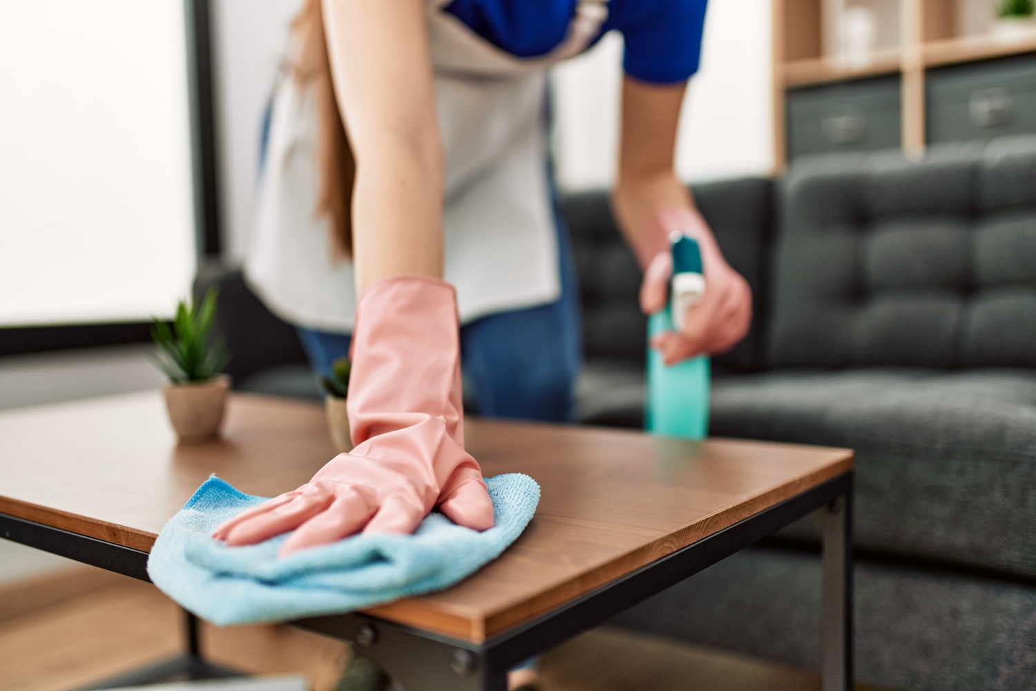 Person cleaning a coffee table with a squirt bottle and a rag while wearing gloves.